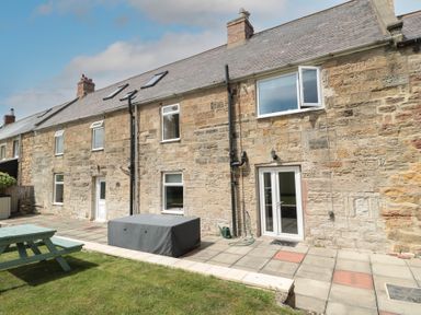 Exterior view of a stone house with multiple windows and doors patio area with covered furniture and green lawn at Changing Tide in Beadnell