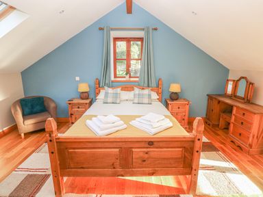 A bedroom with a wooden double bed flanked by two bedside tables with lamps an armchair a wooden dresser with mirrors and a window with curtains at Little Brook Cottage in Laugharne
