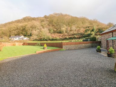 A gravel driveway next to a lawn with potted plants and a stone building near a wooded hill at Little Brook Cottage in Laugharne