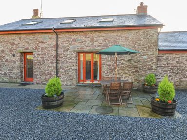 A stone exterior patio with wooden table and chairs under a green umbrella surrounded by planter barrels at Little Brook Cottage in Laugharne