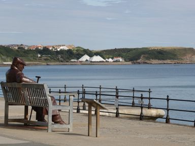 A metal statue of a man sitting on a bench by the water with hills and buildings in the background at Castle View Cabin in Cloughton
