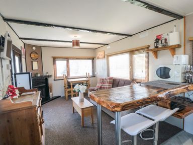 A living room with a wooden table and chairs a brown sofa with pillows a wooden coffee table with flowers a wooden sideboard a microwave and kitchen counter with stools at Castle View Cabin in Cloughton