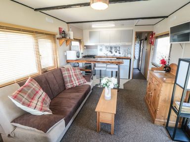 A living room with a sofa and pillows a wooden coffee table with flowers a kitchenette with stools and cooking utensils a wooden cabinet and a wall mounted TV at Castle View Cabin in Cloughton