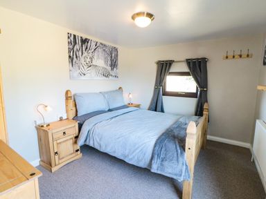 A bedroom with a wooden double bed with gray bedding two wooden nightstands with lamps and a window with gray curtains at Castle View Cabin in Cloughton