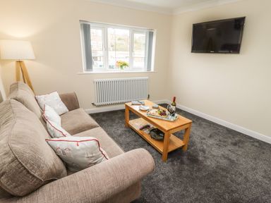 A living room with a beige sofa, wooden coffee table, wall-mounted TV, and window with a flower vase at Station View in Whitby