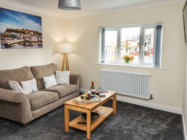 A living room with a sofa and cushions a wooden coffee table with wine and snacks and a window showing a street view at Station View in Whitby