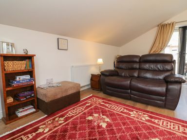 A living room with a brown leather sofa a wooden bookshelf a table lamp and a red patterned rug at Scenic Views in High Bickington