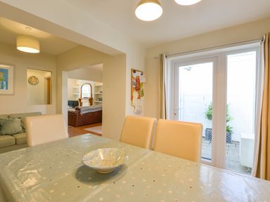 A dining area with a table and chairs next to glass doors and a view into a living room with a sofa at Beachcomber in Padstow