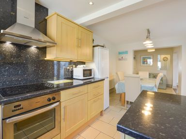 A kitchen with wooden cabinets black countertops a stove microwave and refrigerator next to a dining area with cream chairs and a table with a blue cover at Beachcomber in Padstow