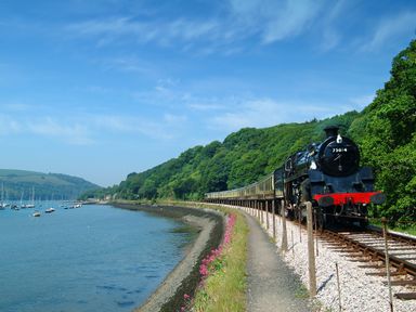 A steam train running along tracks beside a body of water with boats and green hills in the background