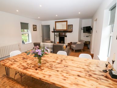 A dining area with a wooden table, white chairs, and a vase of flowers looking into a living room with sofas, armchairs, a fireplace, and a TV at Kealduff Lower in Glenbeigh County Kerry