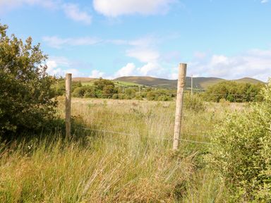 A grassy field enclosed by wire fencing with wooden posts and bushes in the foreground and hills in the background at Kealduff Lower in Glenbeigh County Kerry