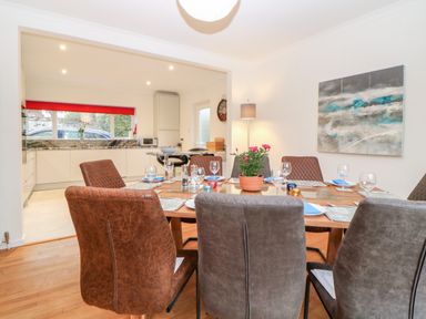 A dining area with a wooden table set with plates wine glasses and a plant in a pot and an open kitchen with white cabinets and a red window blind at Casalmare in Exmouth