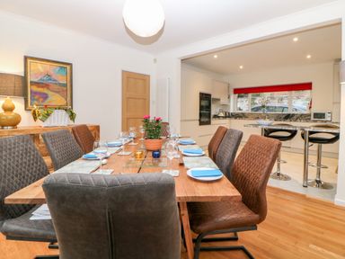 A dining area with a wooden table set with plates and glasses next to a kitchen with bar stools at Casalmare in Exmouth