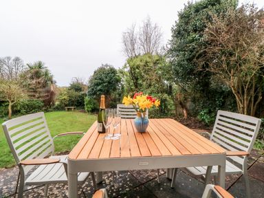 A garden patio with a wooden table and four chairs a vase with flowers a bottle of champagne and three champagne glasses on the table at Casalmare in Exmouth