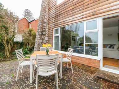 An outdoor patio area with a wooden table, white chairs, flowers in a bottle on the table, next to a house with large windows and an open door at Casalmare in Exmouth