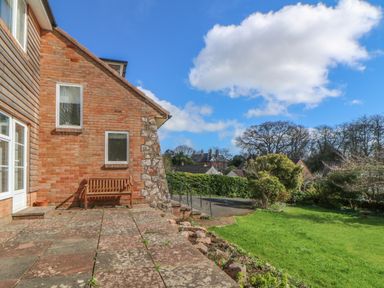 A brick and stone house exterior with a wooden bench on a paved patio next to a green lawn and trees at Casalmare in Exmouth