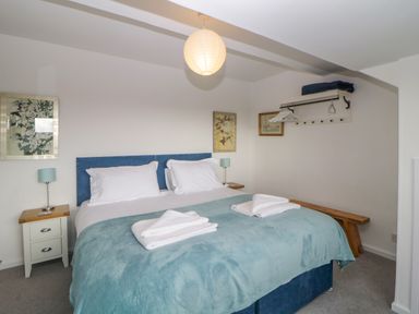 A bedroom with a double bed with blue headboard and teal blanket two bedside tables with lamps wall-mounted clothes rack and wooden bench at Casalmare in Exmouth
