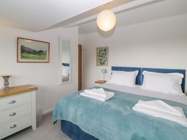A bedroom with a double bed with blue headboard and teal blanket a wooden dresser and wall art at Casalmare in Exmouth