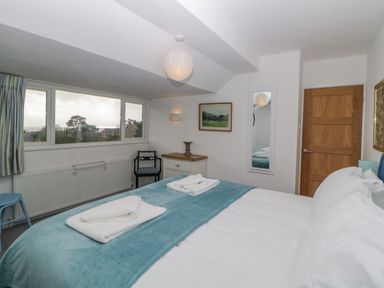 A bedroom with a bed covered in white linens and blue blanket with towels on top a wooden door a wall mirror a nightstand with a vase and a chair near a window at Casalmare in Exmouth