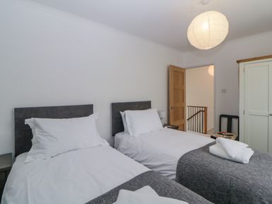 A bedroom with two single beds with white bedding and grey headboards a small nightstand lamp a chair a wardrobe and a wooden door at Casalmare in Exmouth