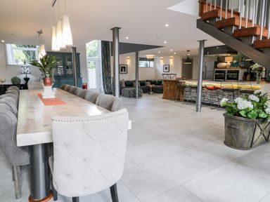 A dining area with a long wooden table and upholstered chairs next to a kitchen with an island and bar stools and a seating area under stairs at Capel Mawr Basement 19 in Llanrug