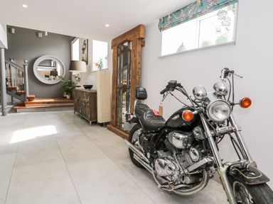 A black motorcycle parked in a hallway with a wooden cabinet, a floor lamp, a round wall mirror, and a large wooden framed mirror at Capel Mawr, Basement 19 in Llanrug