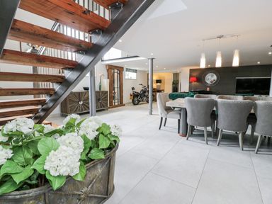 An open living area with metal and wooden staircase white flowers dining table with gray chairs and a motorcycle in the background at Capel Mawr Basement 19 Llanrug