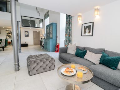 A living area with a gray sofa and pillows a round table with croissants and orange juice a blue cabinet and wall mirror at Capel Mawr Basement 19 in Llanrug