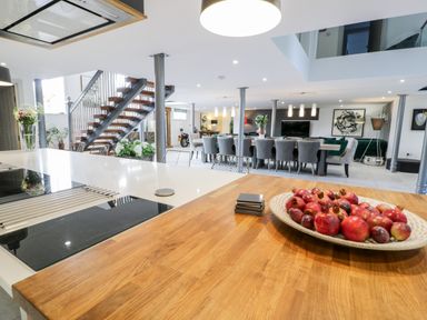 An open kitchen and dining area with a wooden countertop bowl of fruit dining table and chairs a staircase and living area at Capel Mawr Basement 19 in Llanrug