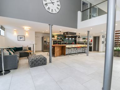 A large open plan kitchen and living area with a gray sectional sofa and a large clock on the wall at Capel Mawr Basement 19 in Llanrug
