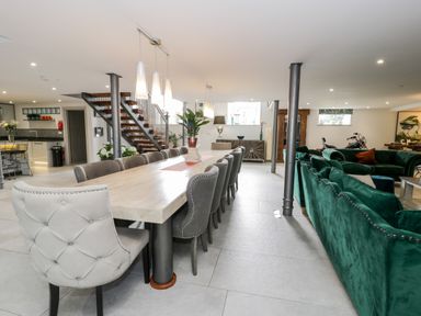 A dining area with a long wooden table surrounded by gray and white chairs next to a living space with green sofas and a staircase in the background at Capel Mawr Basement 19 in Llanrug