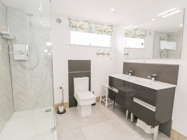 A bathroom with a glass shower enclosure a toilet a double sink vanity large mirror and floral window blinds at Capel Mawr Basement 19 in Llanrug