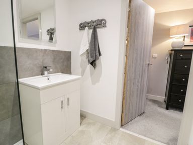 A bathroom with a white sink cabinet grey tiles on the wall towels hanging on hooks and an open door showing a room with a lamp on a dresser at Capel Mawr Basement 19 in Llanrug