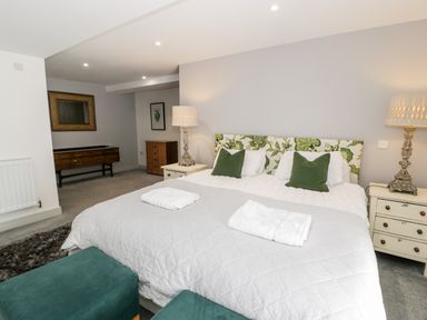 A bedroom with a large bed with floral headboard and green pillows two bedside tables with lamps a green ottoman and wooden dressers at Capel Mawr Basement 19 in Llanrug