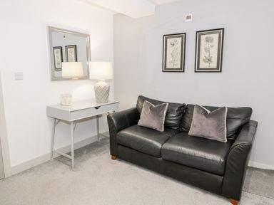A room with a black leather sofa with two cushions a white desk with a lamp and a decorative object a mirror and two framed botanical prints on the wall at Capel Mawr Basement 19 in Llanrug