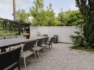 An outdoor patio with a long table and chairs on gravel with trees and a wooden fence at Capel Mawr Basement 19 Llanrug