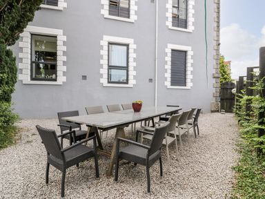 An outdoor dining area with a long table and multiple chairs on gravel next to a gray building at Capel Mawr Basement 19 in Llanrug