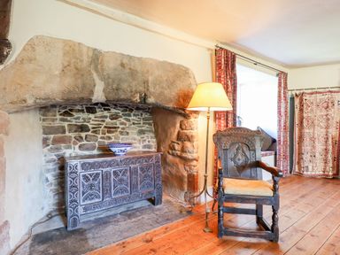 A room with an old stone fireplace wooden carved chest floor lamp wooden chair with cushion and patterned curtains at Aluric's Hall in Porlock