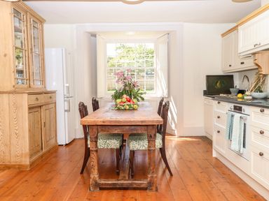 A kitchen with a wooden dining table and chairs a large window and cabinets at Aluric's Hall in Porlock