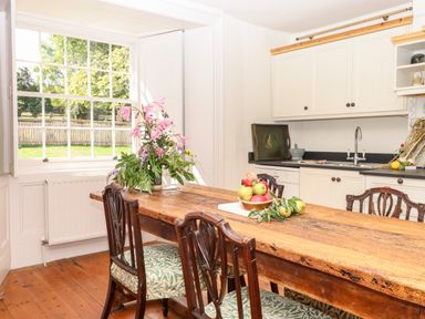 A kitchen with a wooden dining table and chairs a vase with flowers on the table bowl of fruit on the table white cabinets and a sink under the window at Aluric's Hall in Porlock