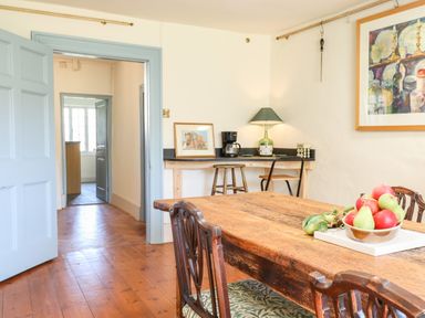 A dining room with a wooden table and chairs a bowl of fruit a side table with a lamp coffee maker framed picture and a painting on the wall at Aluric's Hall in Porlock