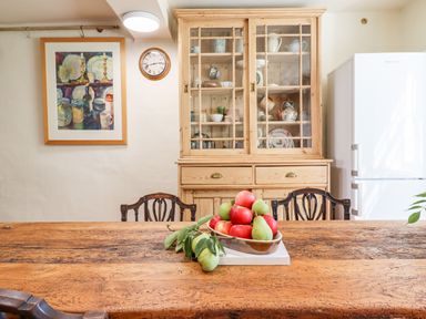 A wooden dining table with a bowl of apples and pears and chairs in a kitchen with a wooden cabinet a clock a framed painting and a white refrigerator at Aluric's Hall in Porlock