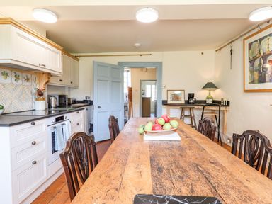 A kitchen with a wooden dining table with fruit bowl and antique chairs next to white cabinets and black countertop at Aluric's Hall in Porlock