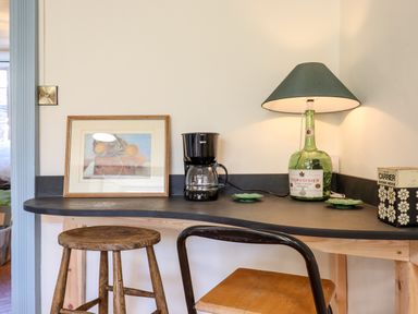 A countertop with a framed picture a coffee maker a lamp made from a bottle and plates and two stools at Aluric's Hall in Porlock