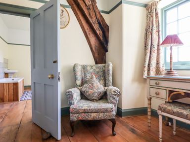 A room with a floral upholstered armchair a wooden desk with a lamp a window and an open door leading to a bathroom at Aluric's Hall in Porlock