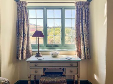 A window with floral curtains above a desk with a lamp and bowl at Aluric's Hall in Porlock