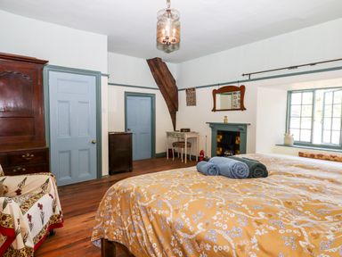 A bedroom with a double bed with yellow floral bedding and rolled towels on top a wooden wardrobe two blue doors a chair with a patterned cover a fireplace with a mirror above a small desk and a window with green trim at Aluric's Hall in Porlock