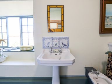 A bathroom with a pedestal sink with decorative tiles and a mirror above a bidet and window with items on the sill at Aluric's Hall in Porlock