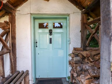 A light blue wooden front door with glass panels surrounded by white walls and stacked firewood at Aluric's Hall in Porlock